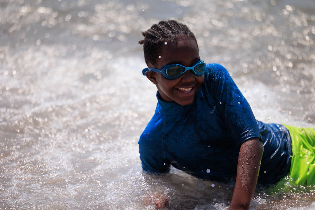 A child wearing a blue rash guard and goggles smiles while playing in shallow water. The background shows the splashing waves.