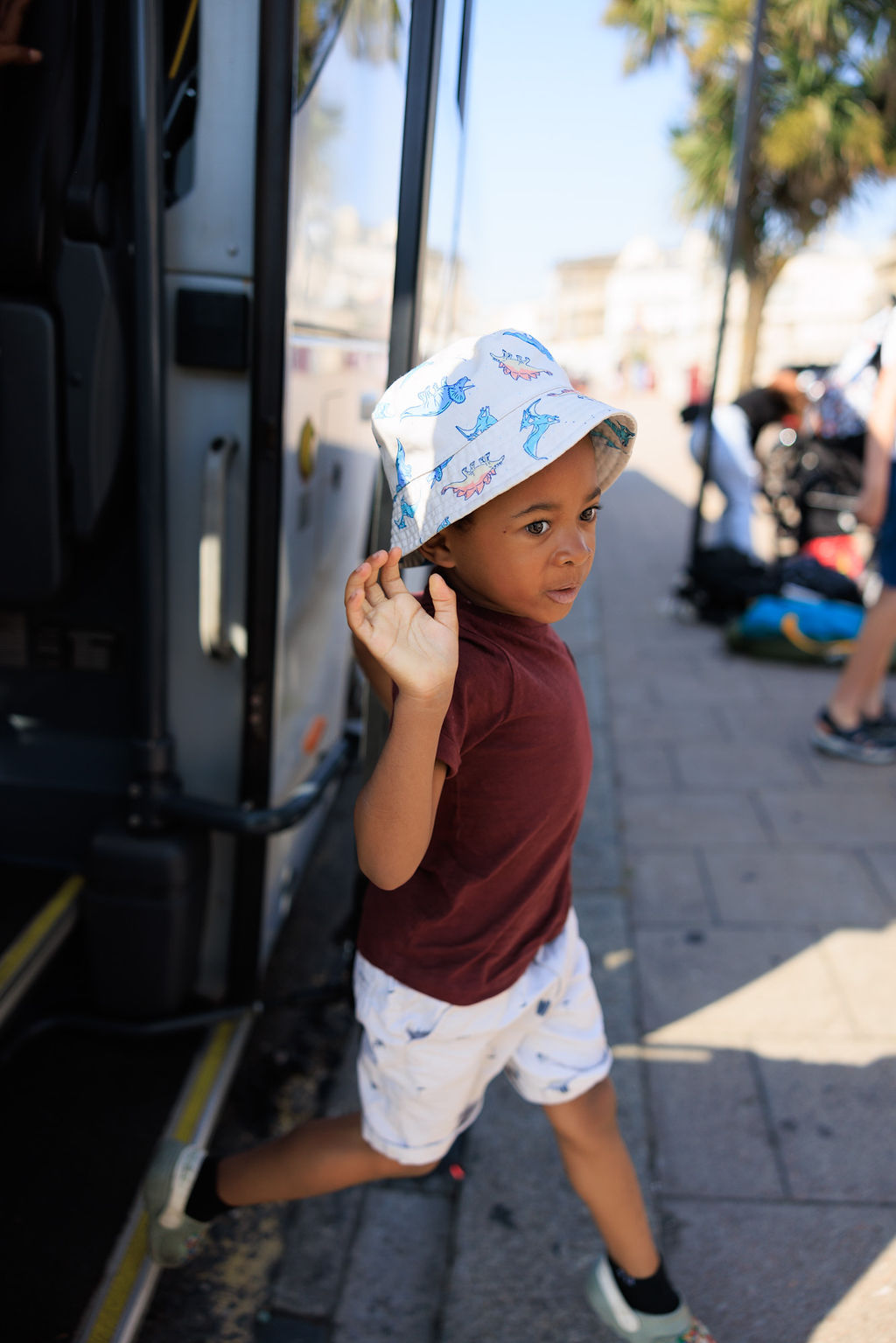 A young child wearing a printed bucket hat, a maroon shirt, and patterned shorts steps off a bus onto a sunlit sidewalk. There are people and palm trees blurred in the background.