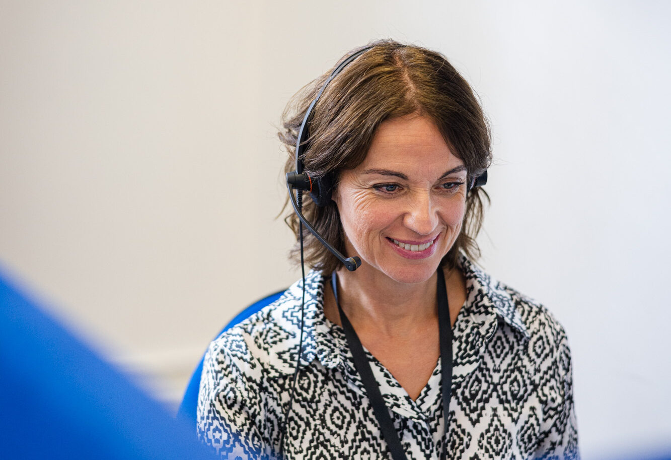 A woman with brown hair is smiling while wearing a headset, seated at a desk. Representing our helpline, she dons a black and white patterned shirt. The softly blurred background suggests an office environment, enhancing the welcoming atmosphere.