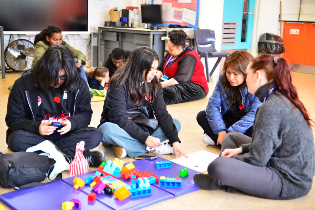 A group of adults and children are seated on the floor in a classroom, engaging in an activity with colorful building blocks. Some are focused on instructions, while children play nearby. The room is bright and casual.