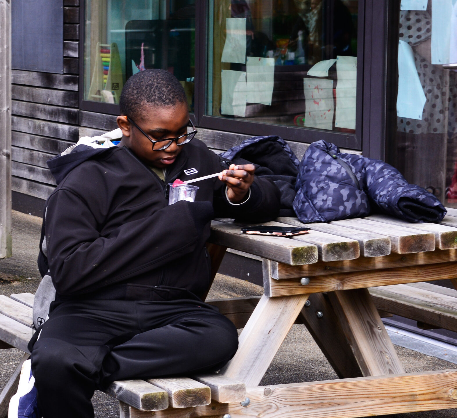 A boy in a black tracksuit and sneakers is sitting on a wooden picnic table outdoors, focused on reading a book or paper. Coats and a backpack are on the table. Reflections in a window show other people indoors.