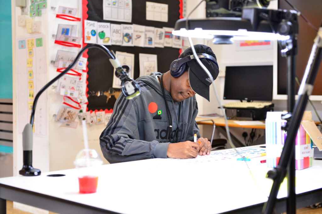 A person wearing headphones and a visor concentrates on coloring a page at a well-lit desk. The space is filled with educational materials and lighting equipment, creating a focused and creative learning environment.