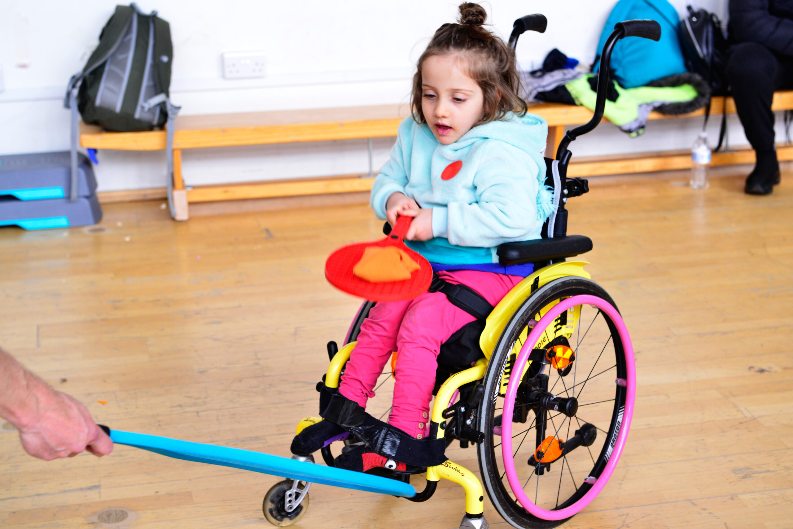 A young child in a wheelchair holds an orange paddle, engaging in a game with a blue foam stick held by another person. The child is wearing a blue jacket and pink pants. The setting appears to be an indoor recreational space.