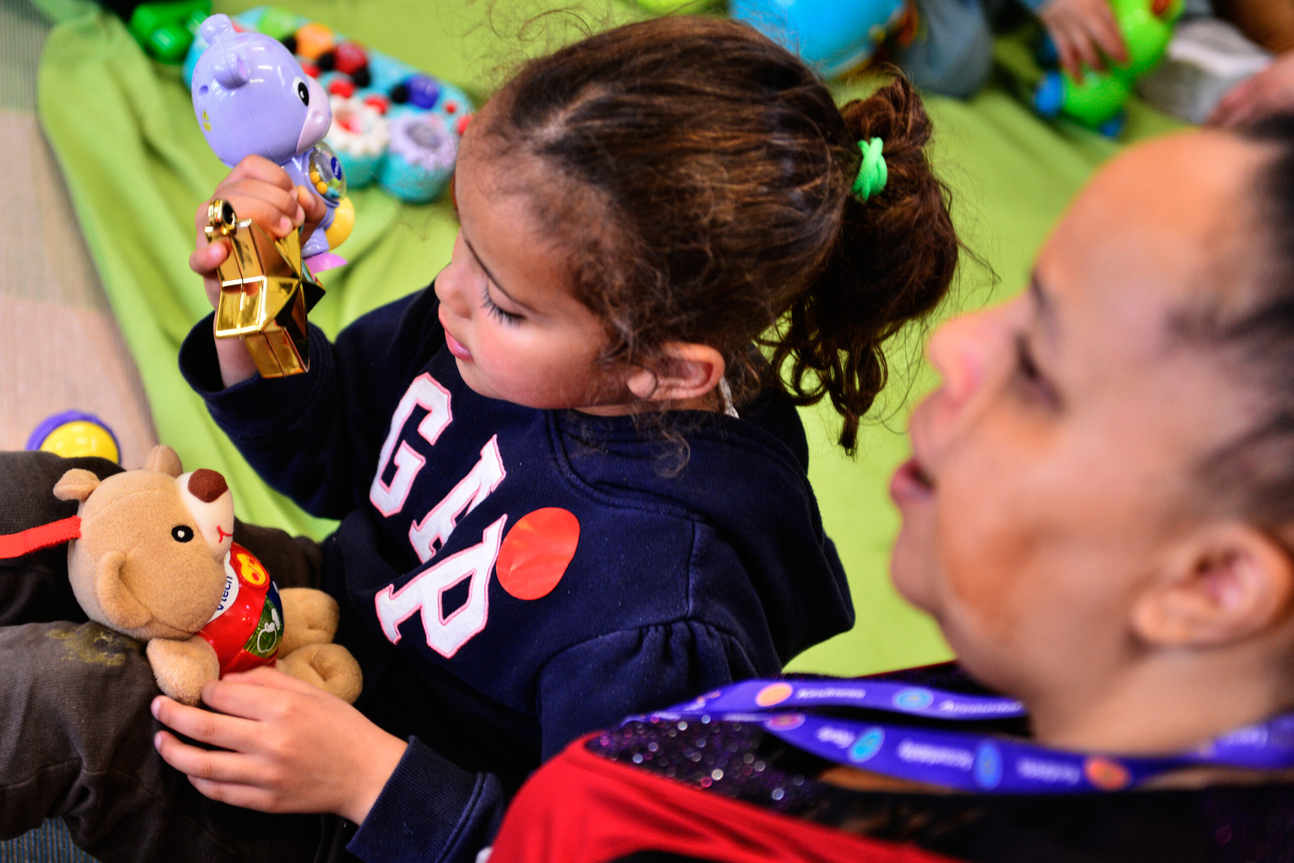 A young girl with curly hair sits next to an adult, holding a toy bear in each hand. They are on a green mat, with various colorful toys scattered around. The girl's navy hoodie has “GAP” written on it. The adult has a lanyard around their neck.