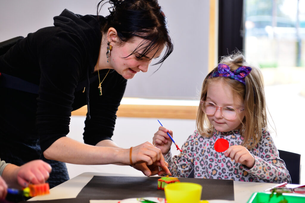 A woman and a young girl are painting together at a table. The woman is guiding the girl, who holds a brush and wears glasses and a floral top. Art supplies are scattered around, and they seem focused and happy.