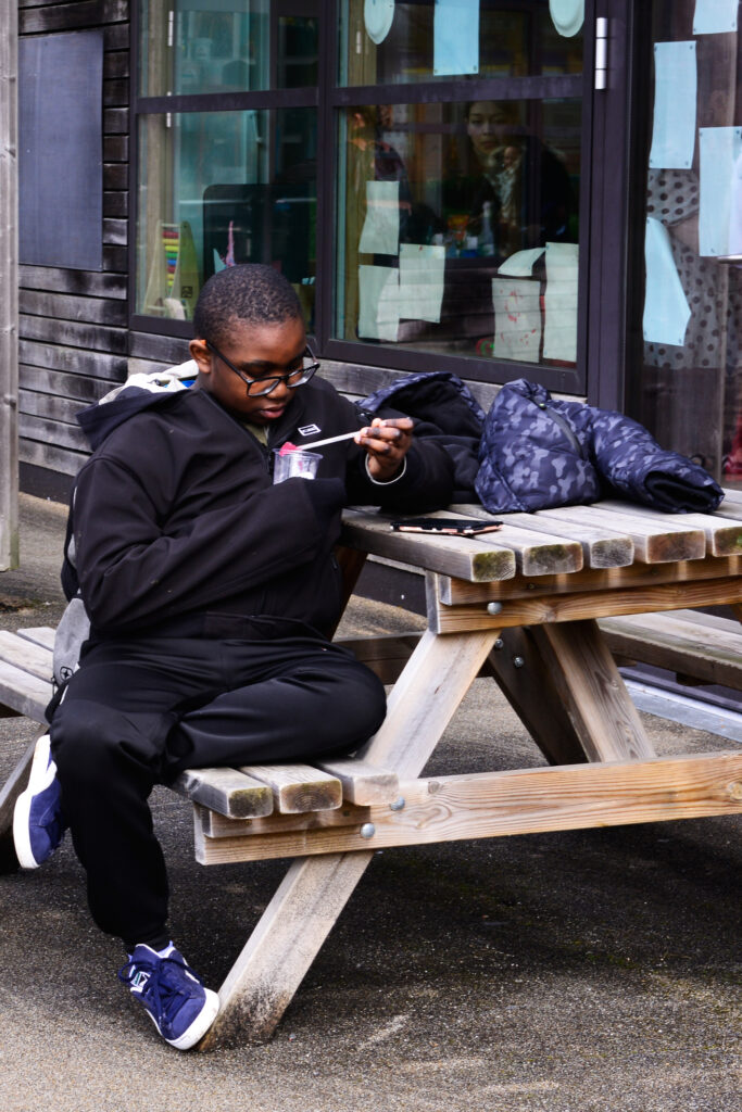 Boy sitting at a wooden picnic bench, engrossed in reading a booklet. He's wearing a black tracksuit and blue sneakers, with a jacket and bag beside him. A window with displays is in the background.