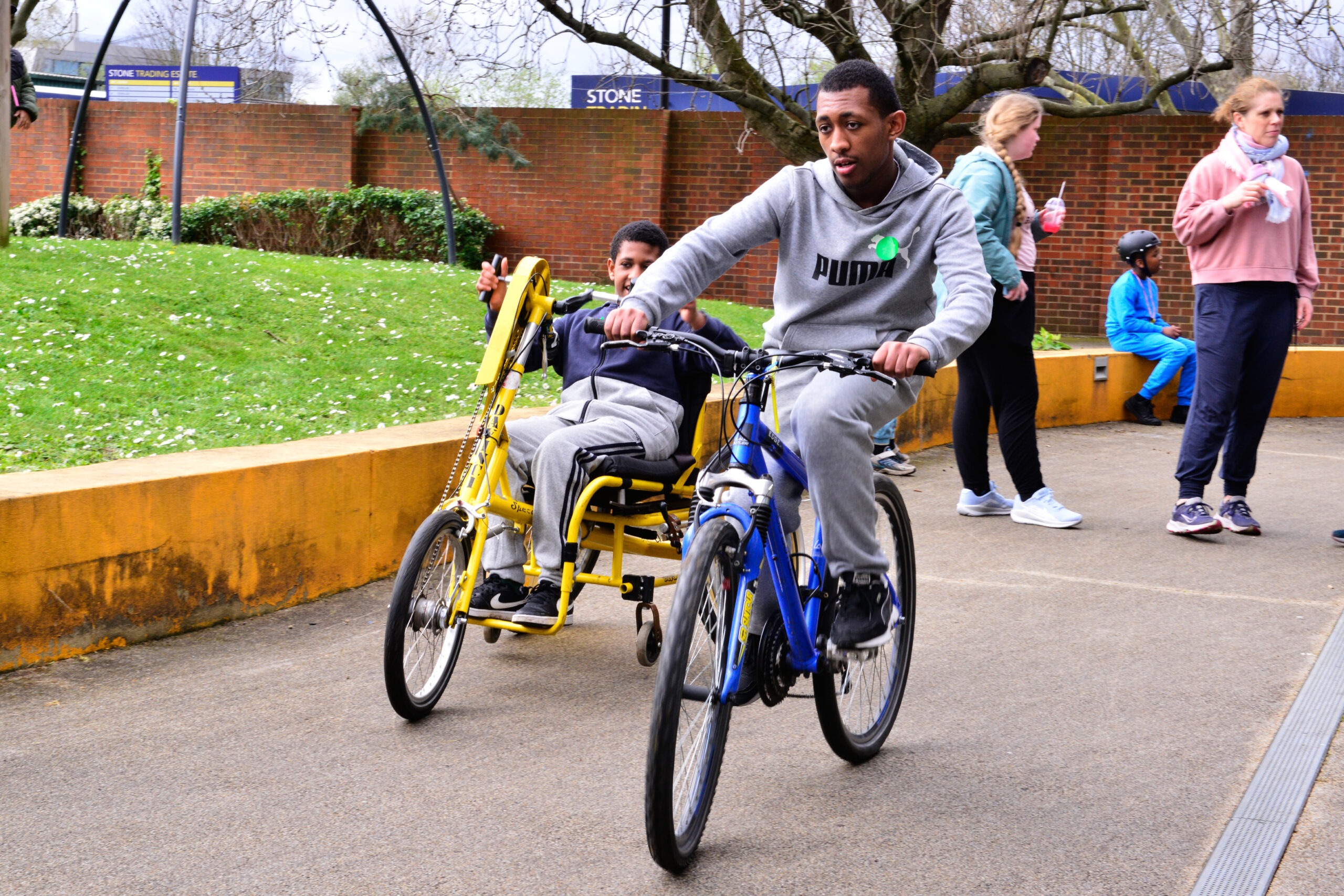 A man rides a blue bicycle while guiding a child seated on a connected yellow tricycle. They are outdoors, surrounded by grass, a brick wall, and other people walking or standing nearby.