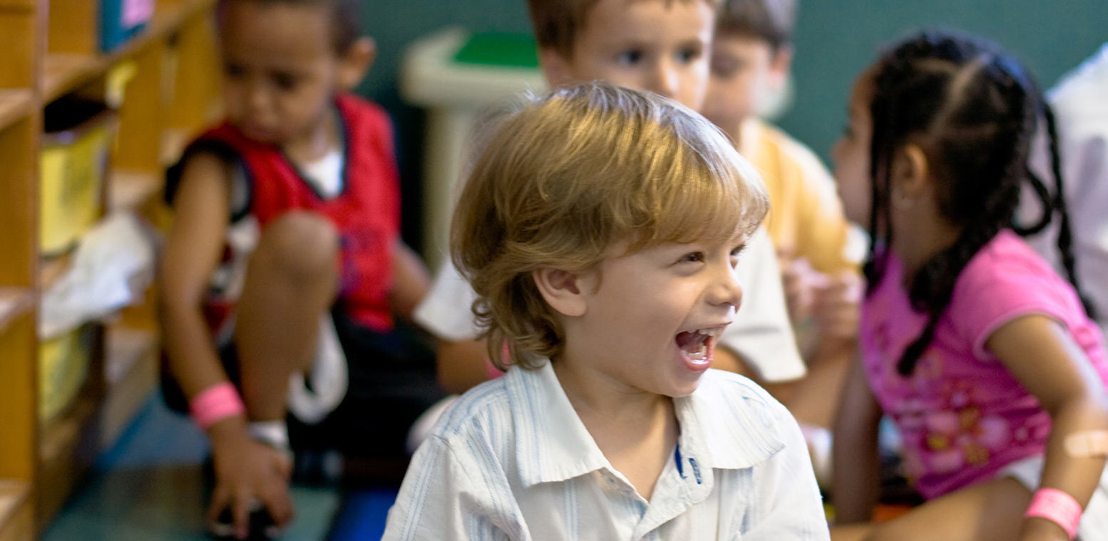 Children sitting in a library setting - one child is in the foreground in focus, smiling off to the side; children are gathered behind in soft focus.