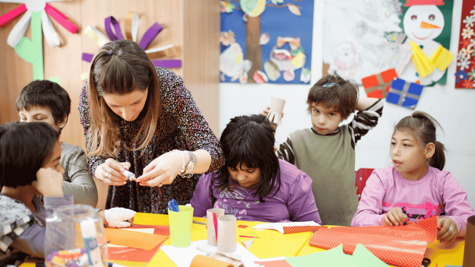 A teacher helps students with a craft activity at a table covered with colorful paper. Children surround the teacher, some focused on the project, while others look on. The wall displays kid artwork, including a snowman and a tree.