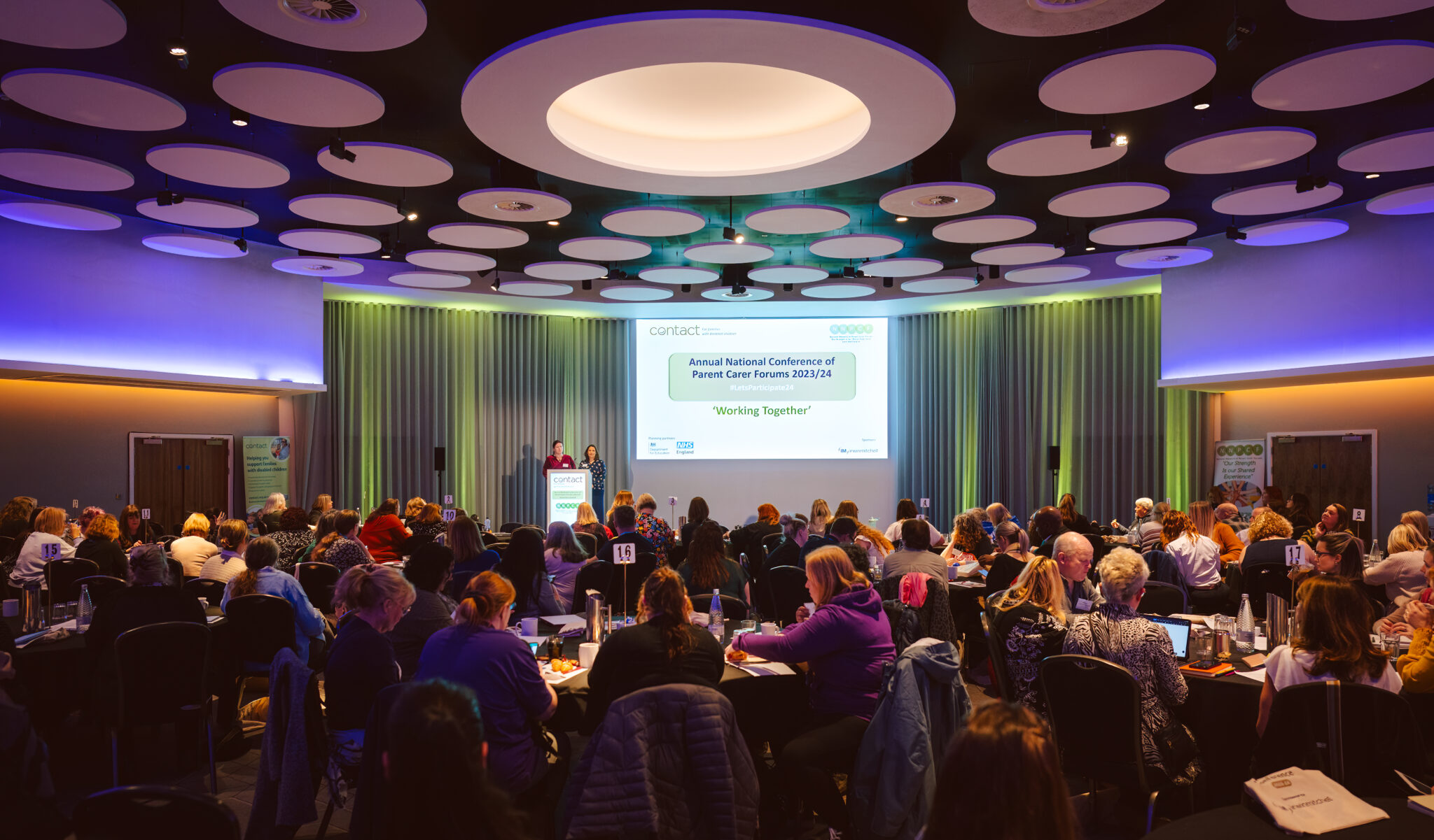 Two people presenting on stage in front of a conference room full of attendees set around round tables.
