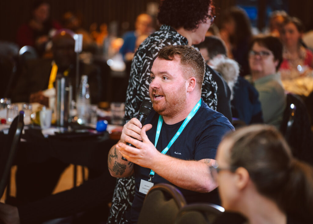 A person speaking into a microphone at an indoor event, surrounded by seated attendees. The speaker wears a lanyard, and the room has a warm, engaging atmosphere. Other event materials and people are visible in the background.