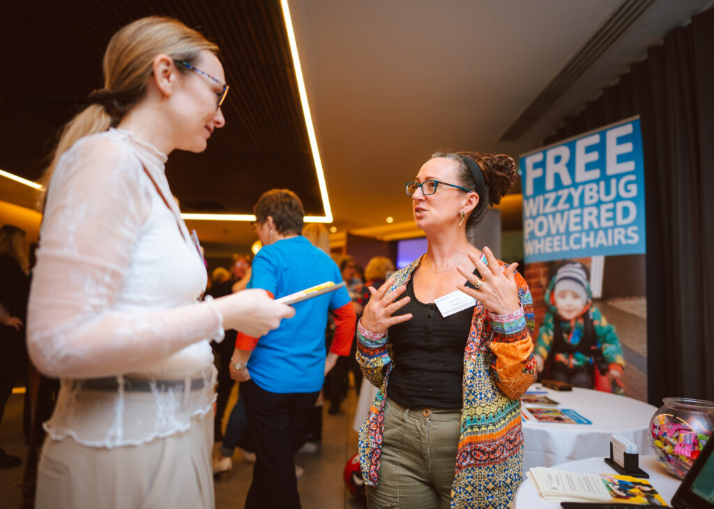 Two women are engaged in conversation at an indoor event. One is holding a tablet, and the other is gesturing with her hands. A sign in the background reads,