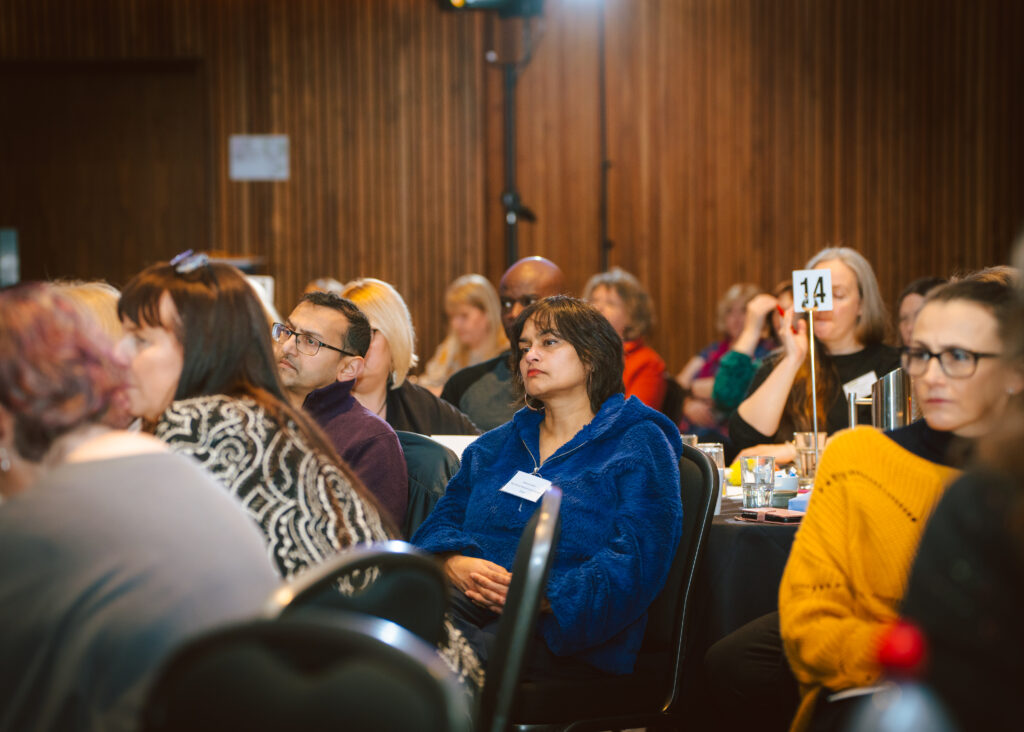 A group of people seated and attentively listening at an indoor event. They are in chairs, with drinks and programs on the tables. A table number reading 