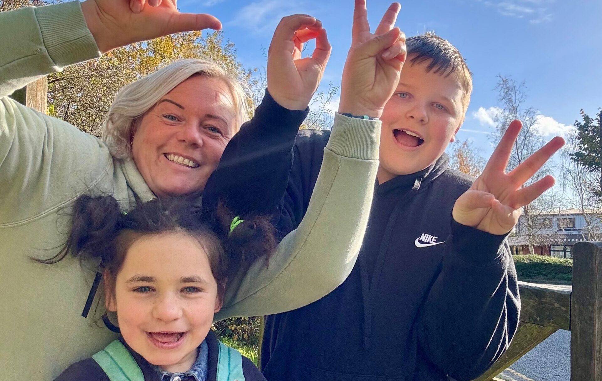 A woman with and two children stand outside, smiling and making hand signs. The older child wears a black hoodie, the younger one has pigtails, all under a clear blue sky with trees in the background.