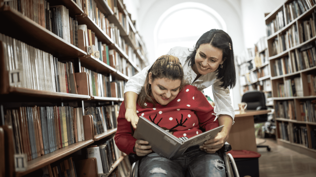 Disabled teenager in wheelchair reading a book between library shelves, with a non-disabled peer looking over their shoulder.