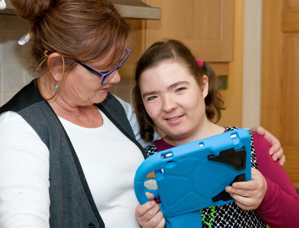 A child holding a tablet smiling at the camera, with her parent standing next to her with her arm around her.