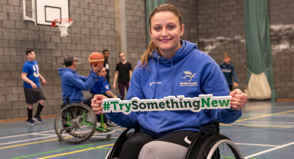 A young woman in a wheelchair holds a sign saying 'TrySomethingNew' in front of a group of wheelchair basketball players on a court.