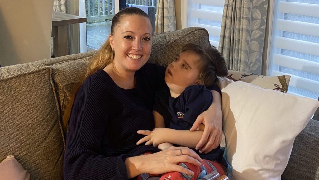 A mum hugs her son on a sofa in front of a window.