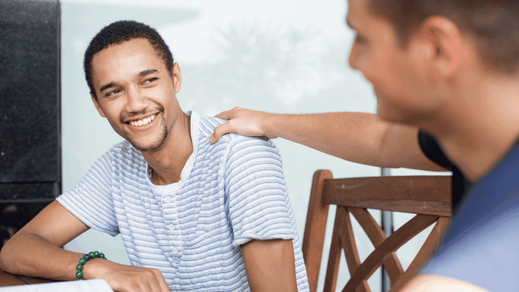A man in a blue t shirt puts his arm on the shoulder of his friend, a smiling man in a white t shirt.