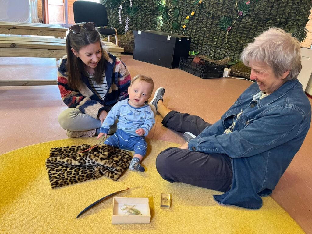 Two adults sitting around and smiling at a young child on a yellow rug. The child sits in front of an insect museum artifact.