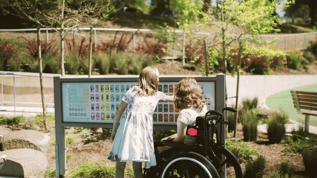 Two children in the park looking at some signage on a sunny day. One child is in a wheelchair, the other is standing pointing at the sign.