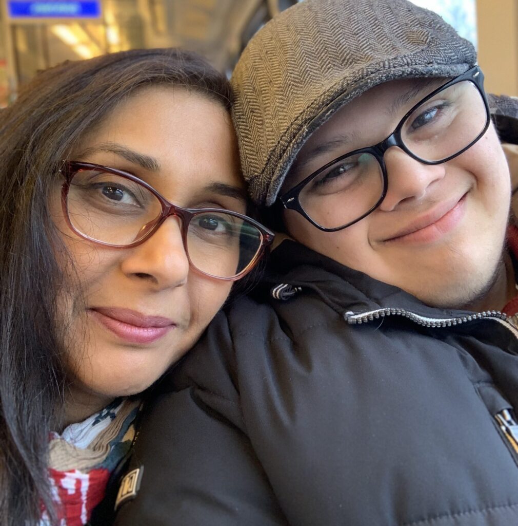 A woman and a young person with glasses and a hat are sitting close together, smiling warmly at the camera whilst riding disabled school transport in what appears to be a public transport setting.