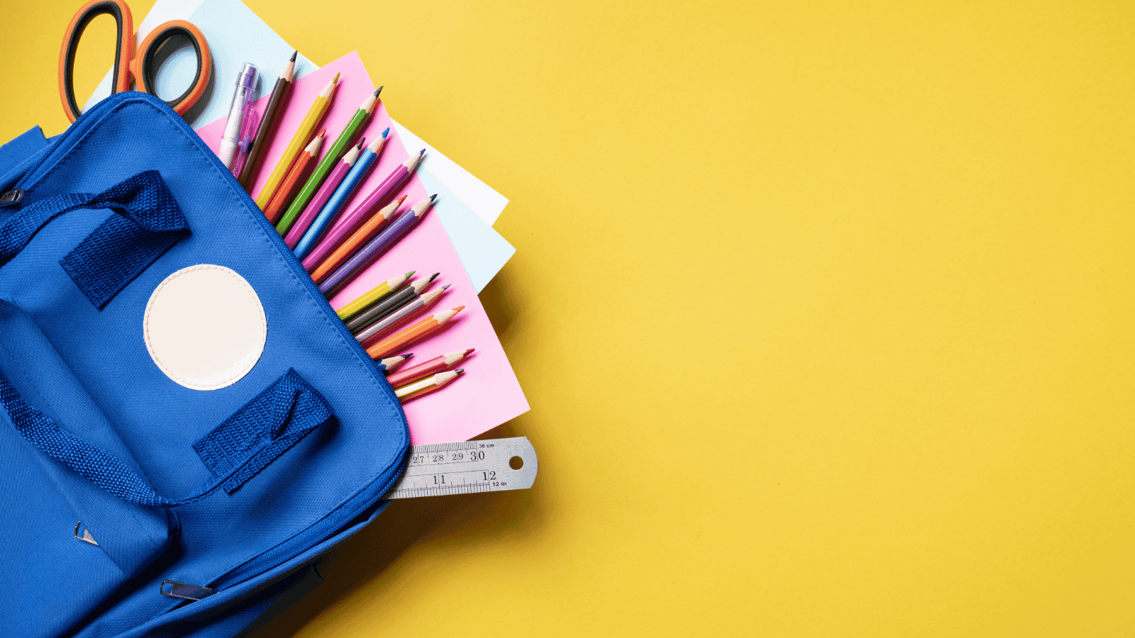 An open blue backpack on a yellow background. Poking out of the bag are colour pencils splayed in a fan shape, some scissors and paper.