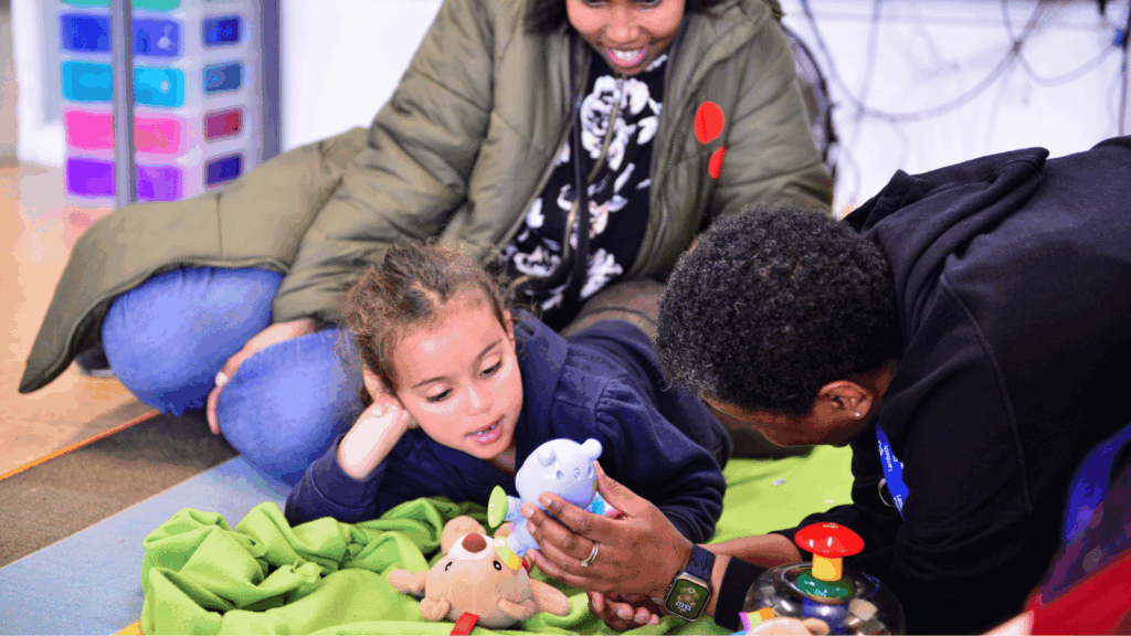 A young child lies on a green blanket, engaged with a blue toy held by an adult. Another adult watches, smiling. 