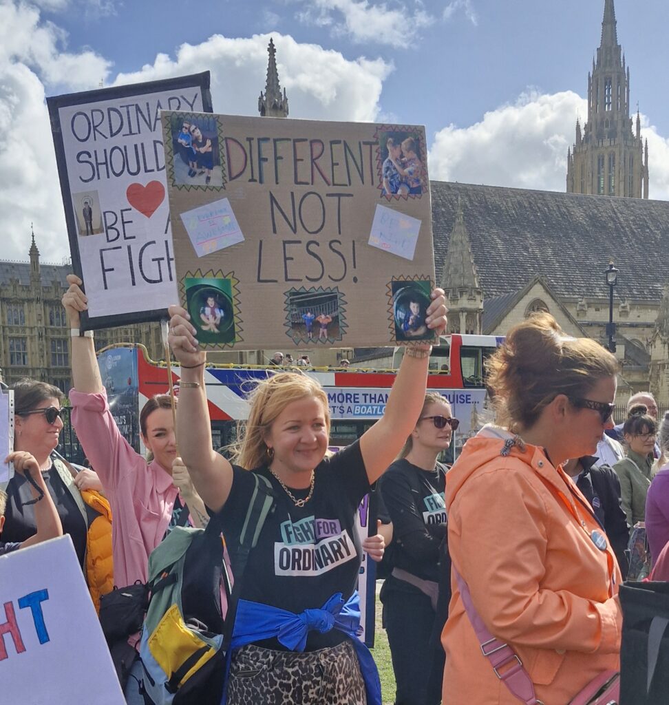 Woman wear a fight for ordinary t-shirt stands outside Parliament holding a banner above her head. The banner features the words 'Different not less''