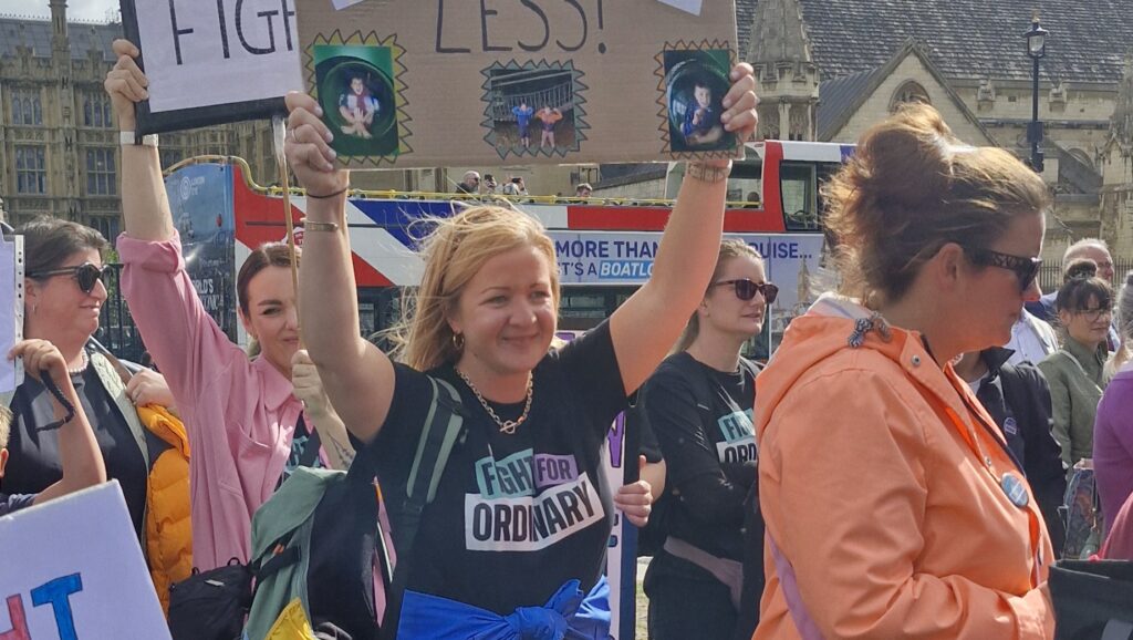 Woman wear a fight for ordinary t-shirt stands outside Parliament holding a banner above her head. The banner features the words 'Different not less''