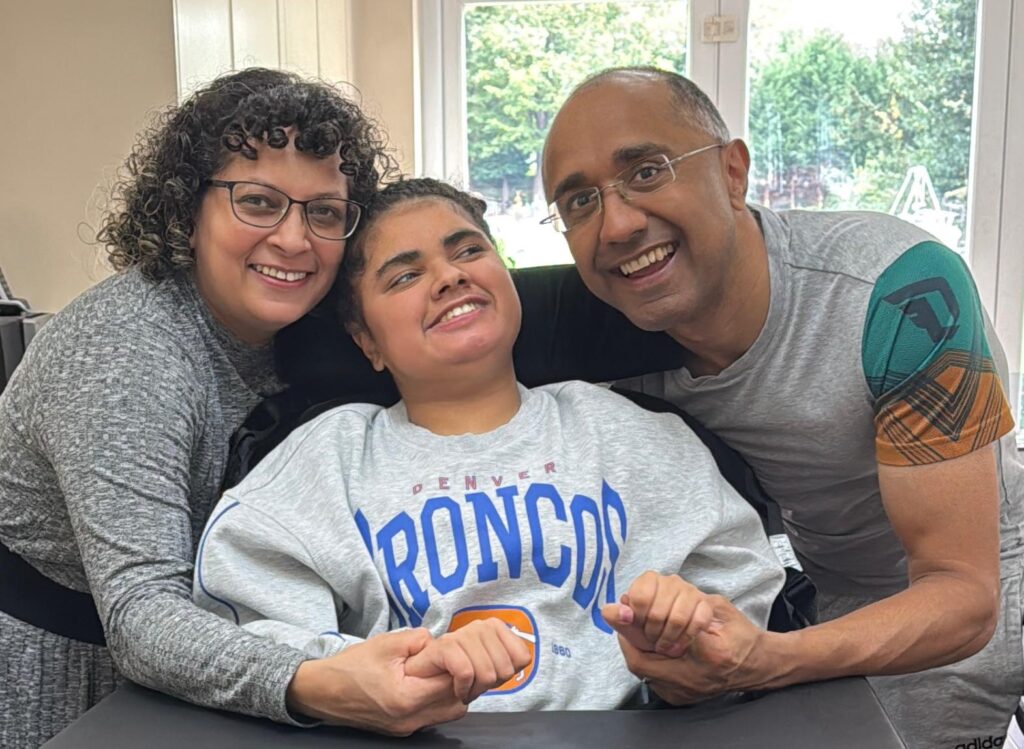 Two parents either side of their teenage daughter, holding her hands. They are all smiling into the camera.