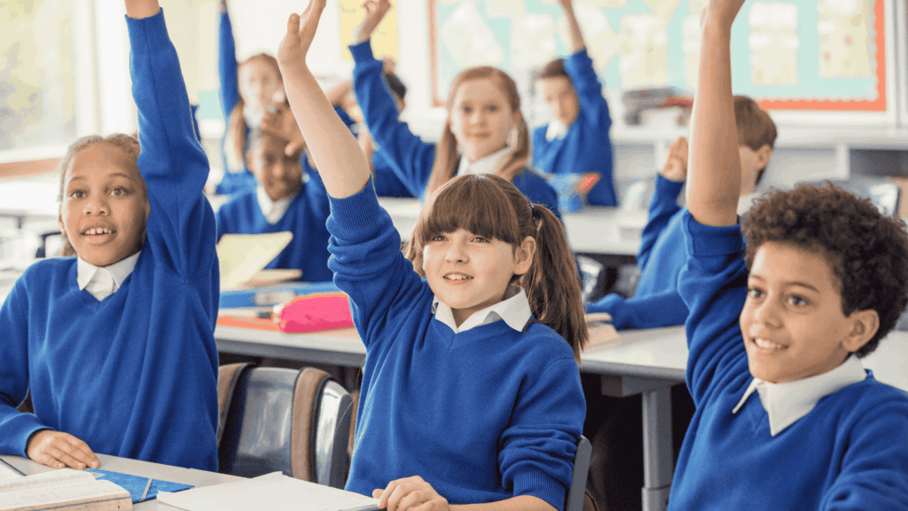 A group of children in a classroom with their arms raised. They are all wearing blue jumper school uniform.