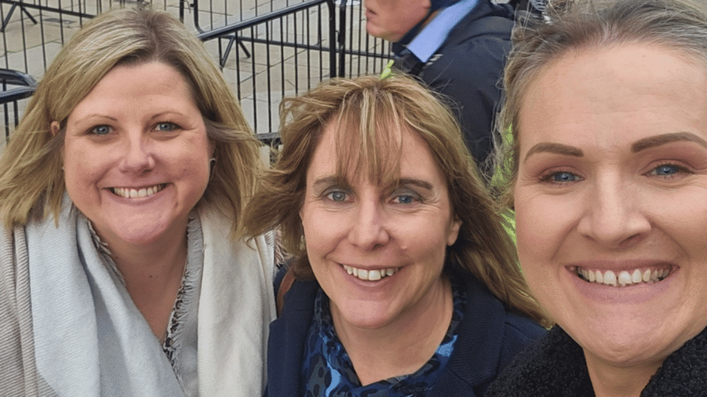 Three women from '3 Mums 1 Mission ARFID' podcast smiling at the camera outdoors with metal railings in the background.