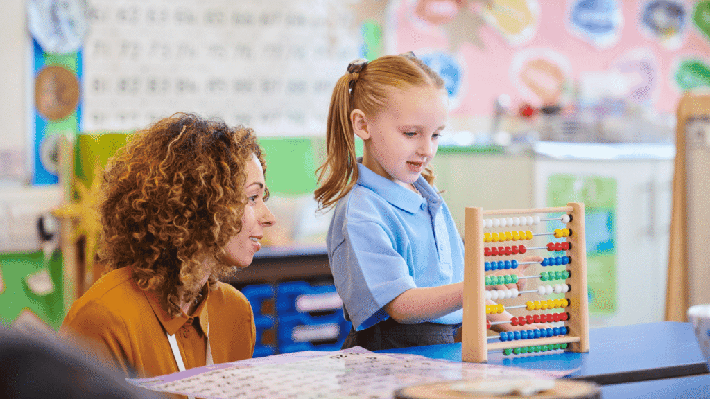 A young pupil in a blue polo shirt using an abacus as a teacher crouches next to her looking on.