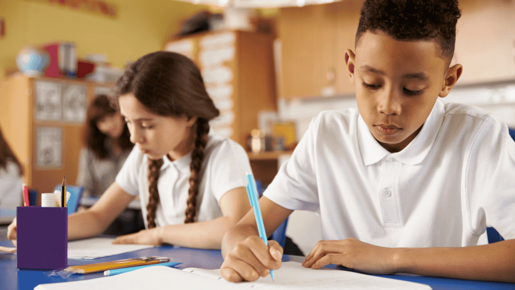Two students sat at a desk in white polo shirts writing on paper pads