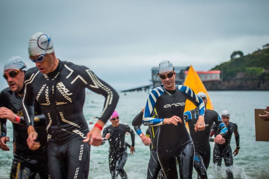 A group of athletes wearing wetsuits and swim caps run out of the water during a triathlon, with a yellow buoy and shoreline in the background under cloudy skies.