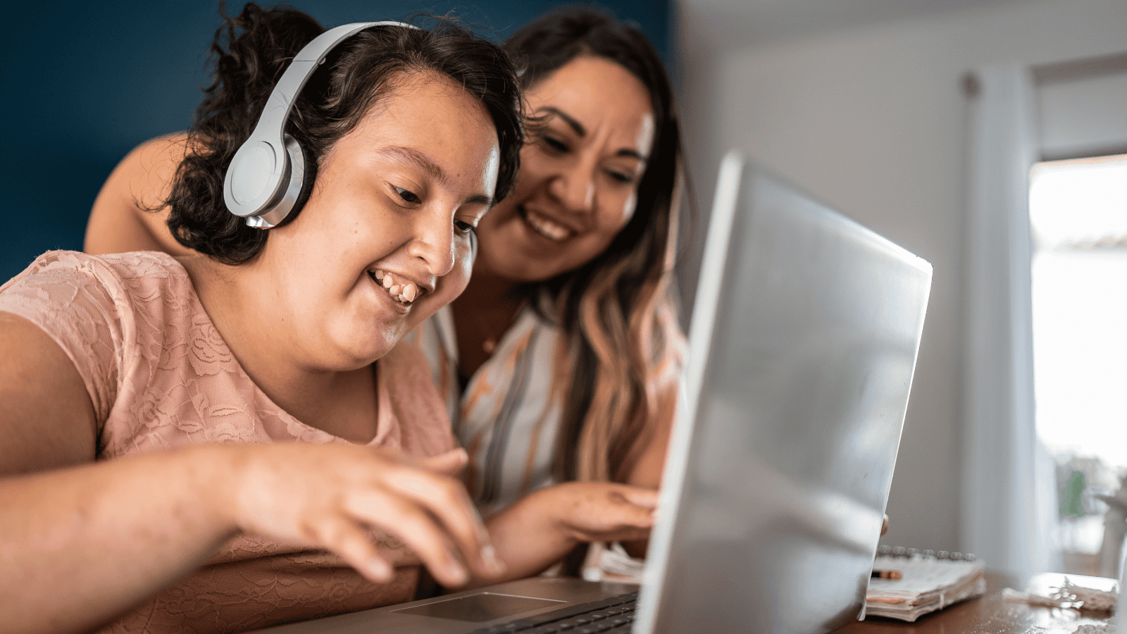 A girl wearing headphones smiles whilst using a laptop, with a woman beside her also smiling and looking at the screen. They appear to be enjoying the activity together in a home setting.