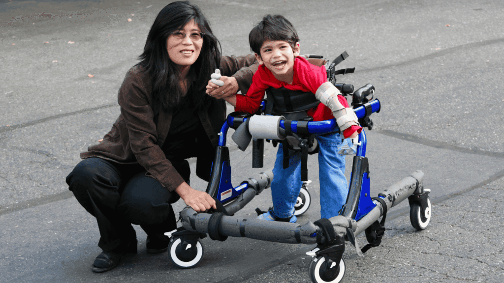 A woman kneels next to a smiling young boy using a blue and grey walker outdoors on a paved surface. The boy wears a red shirt and the woman has long dark hair and glasses.
