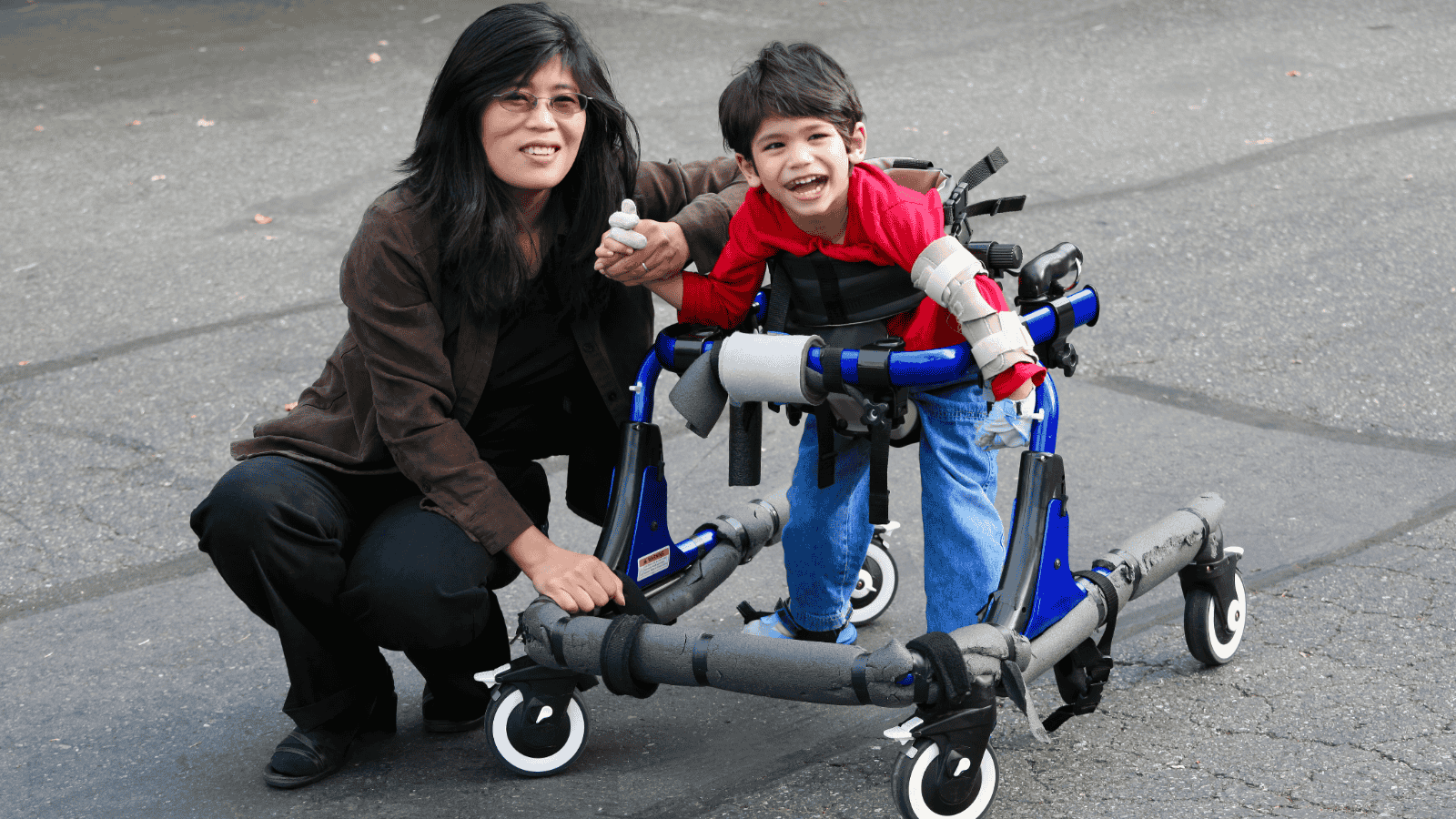 A woman kneels next to a smiling young boy using a blue and grey walker outdoors on a paved surface. The boy wears a red shirt and the woman has long dark hair and glasses.