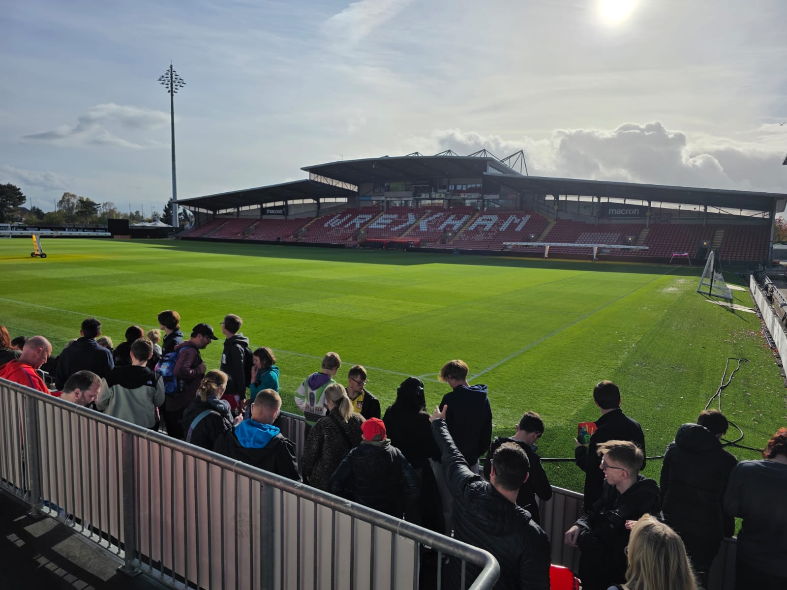A group of people gather near a railing at a football stadium with green grass and red seats. The stadium, under a sunny sky, has "WREXHAM" written across the main stand.