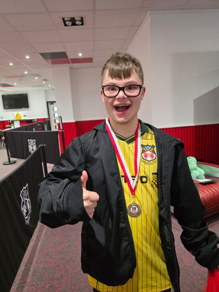 A young man wearing glasses, a yellow football shirt, and a black jacket smiles and gives a thumbs up. He is wearing a medal round his neck and standing indoors in a sports centre.