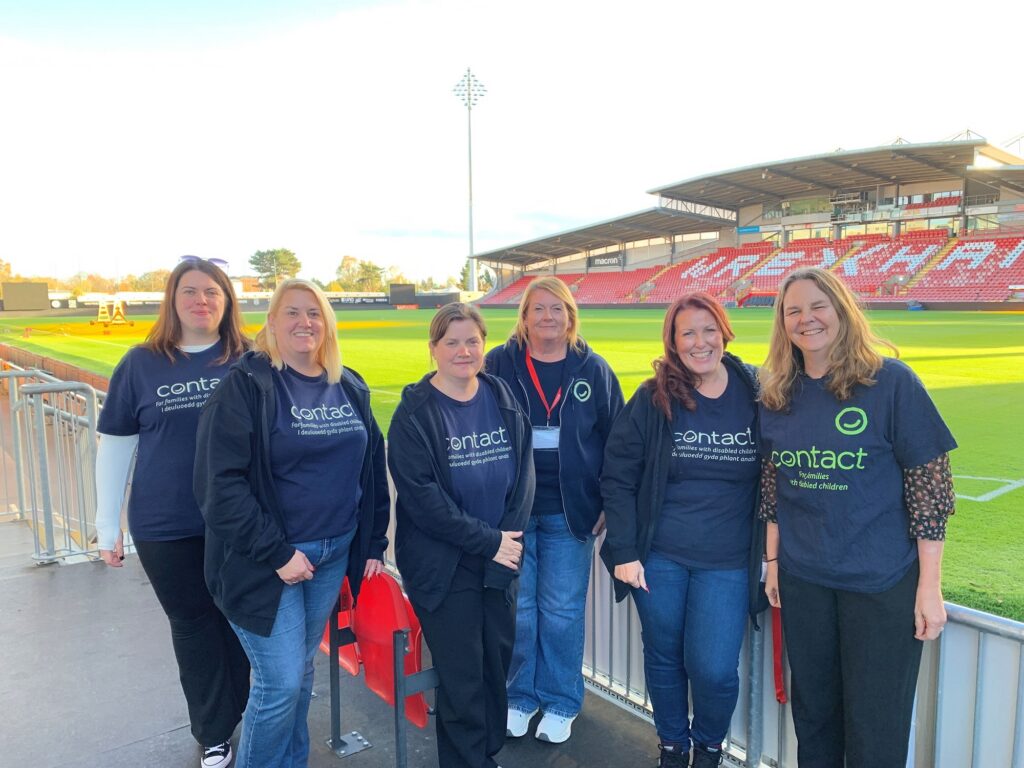 Six women stand side by side at a football stadium, all wearing navy blue "Contact" T-shirts. The stadium seats and pitch are visible in the background under a partly cloudy sky.