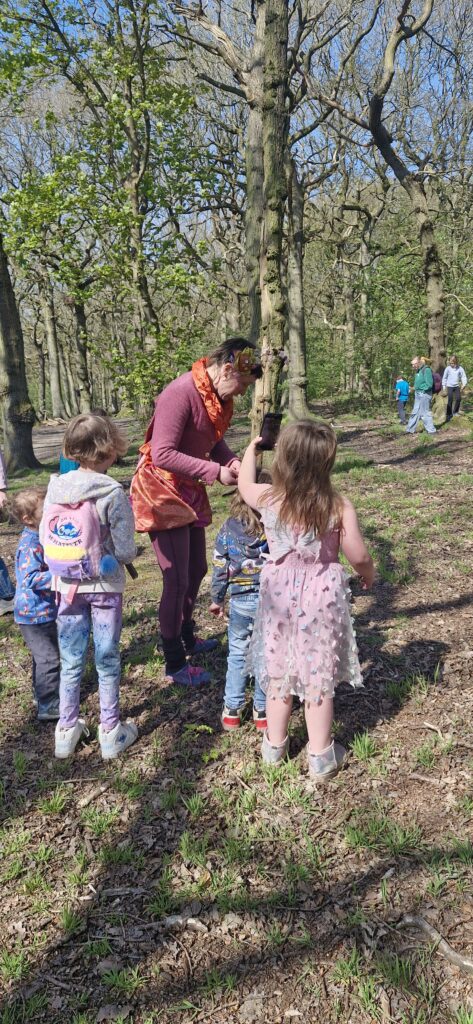 A group of children and an adult stand in a sunlit, open woodland, surrounded by trees. The adult hands something to a child, while other children with rucksacks watch. More people walk in the background among the trees.