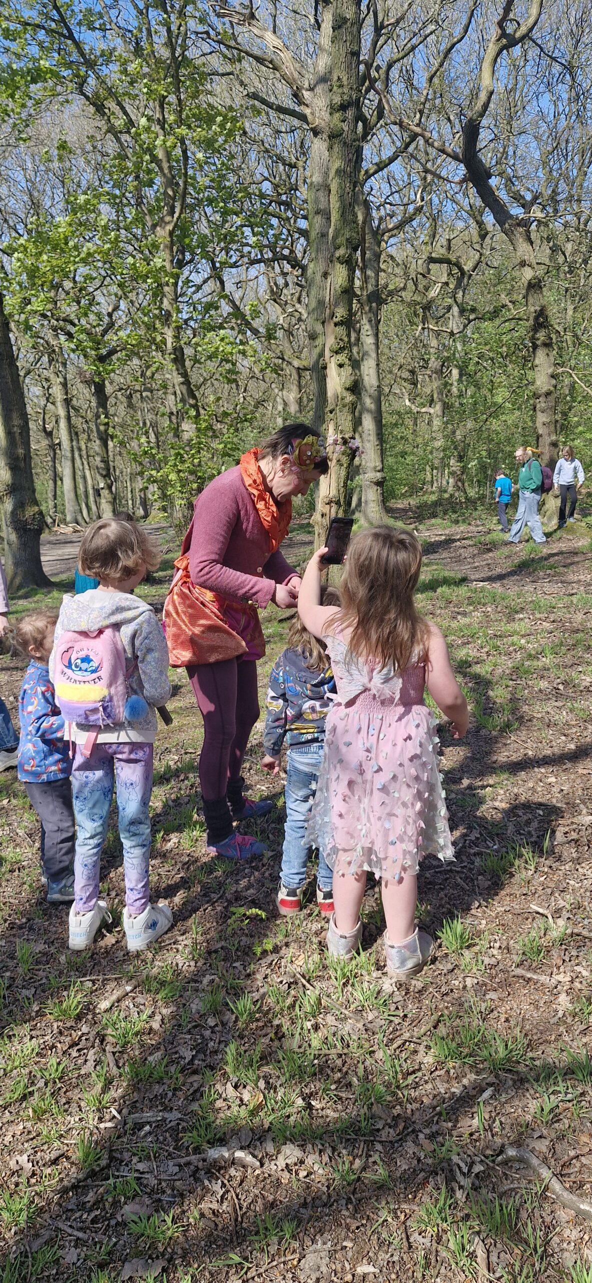 A group of children and an adult stand in a sunlit, open woodland, surrounded by trees. The adult hands something to a child, while other children with rucksacks watch. More people walk in the background among the trees.