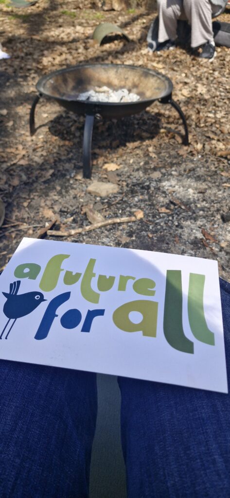 A person holding a sign that reads “a future for all” with a bird graphic sits outdoors near a firepit on the leaf-covered ground of a free woodland. In the background, two people’s legs are visible.