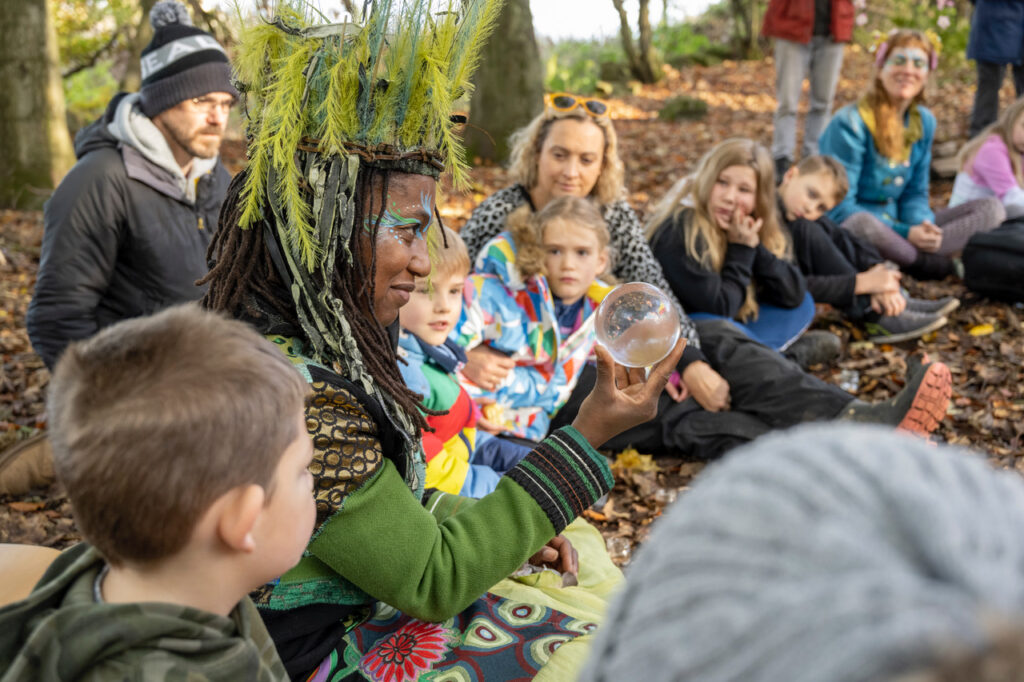 A performer in colourful, nature-themed attire and a feathered headdress holds a crystal ball, entertaining a group of children and adults sitting on fallen leaves in a forest clearing.