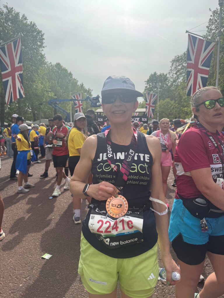 A smiling runner wearing sunglasses, a hat, and a race number (22416) holds a finisher’s medal. Other runners and large UK flags are visible in the background on a sunny day.