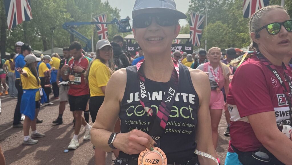 A smiling runner wearing sunglasses, a sunhat, and a medal poses after a race, surrounded by other participants. Union Jack flags are visible in the background, suggesting a UK event. The runner's bib reads 