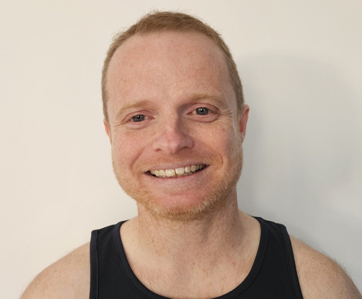 A smiling man with short fair hair and a trimmed beard wears a black vest with the word "contact" in yellow text on the front, standing against a plain white background.