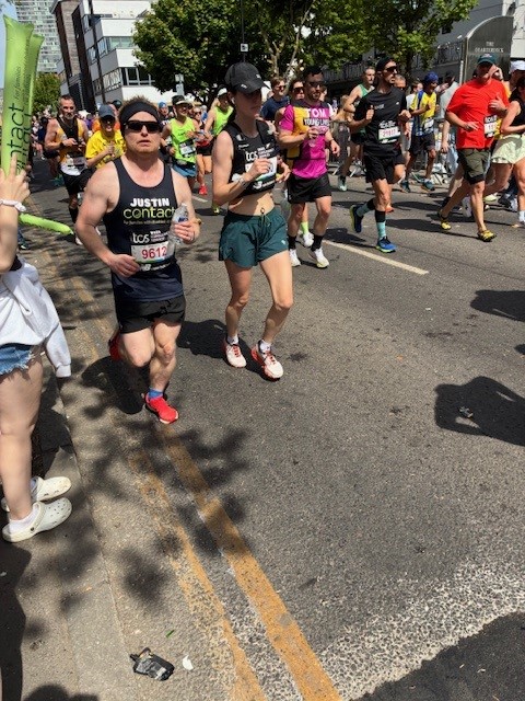 Runners participate in a road race on a sunny day, surrounded by cheering spectators and trees. The street is crowded with people in sportswear and race numbers.
